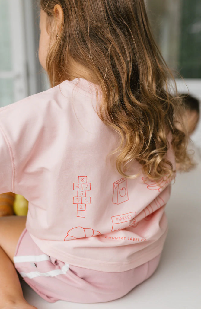 Back view of a child sitting cross-legged, highlighting the fun hand-drawn graphic illustrations in red on the back of the Happy Days Tee.