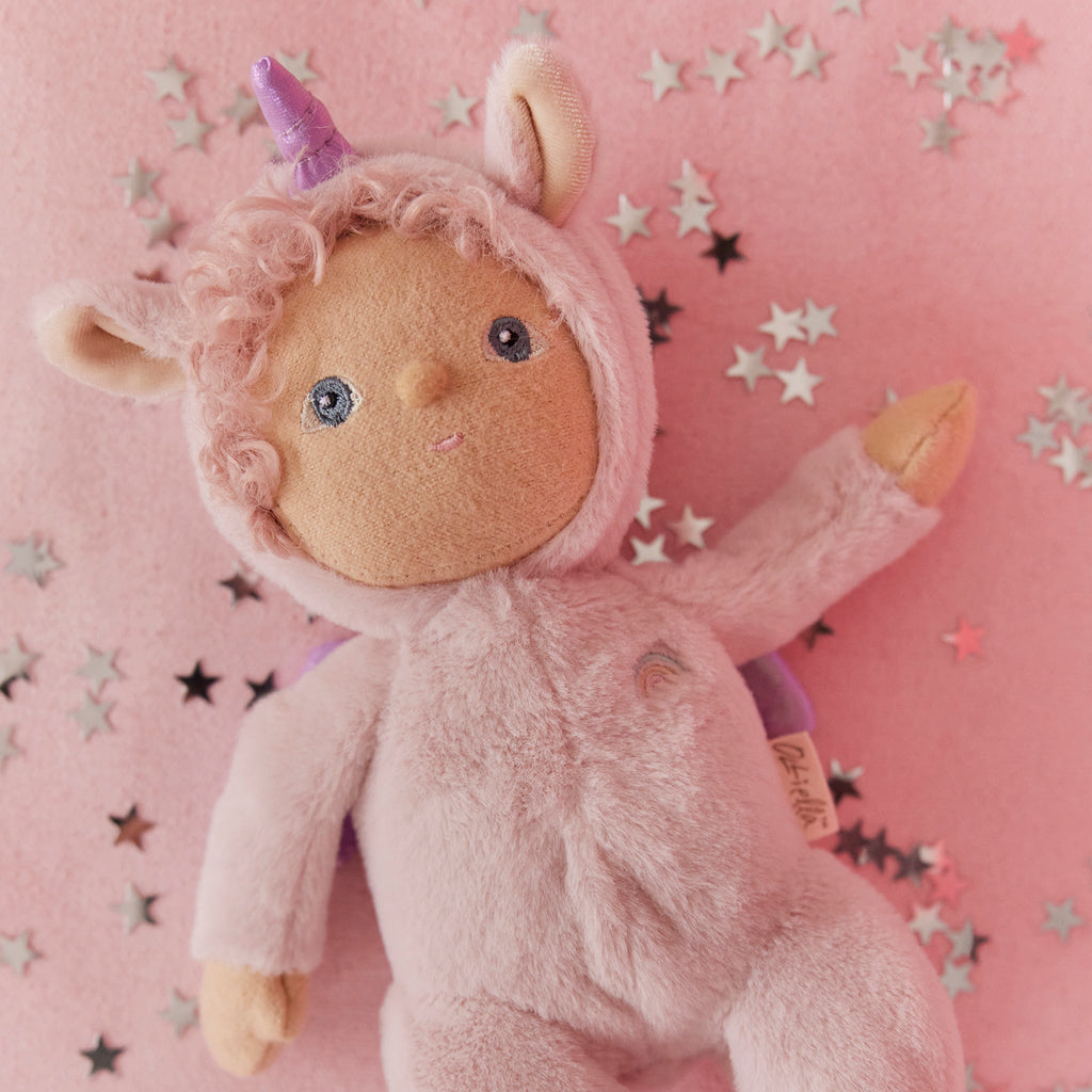 Close-up of Olli Ella Dinky Dinkum Doll Una Unicorn lying on a pink background scattered with silver star confetti, wearing a plush pink unicorn onesie with lilac horn, curly hair, and embroidered rainbow detail.