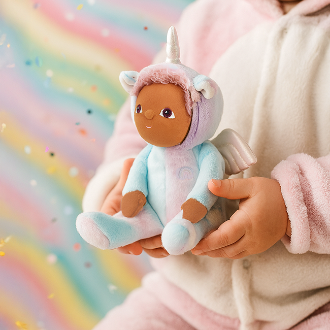 Uli Unicorn Doll against rainbow backdrop – Plush Dinkum unicorn doll being held by a child on a pastel rainbow background with confetti.
