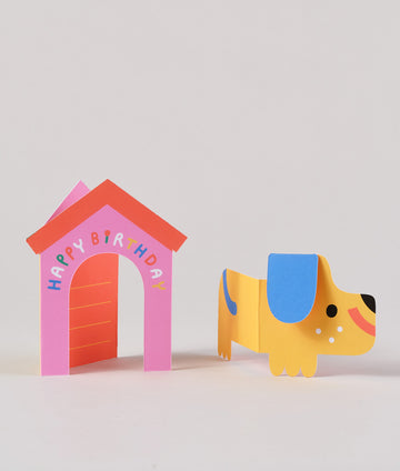 Fold-out birthday card with a yellow dog popping out of a pink doghouse that says “HAPPY BIRTHDAY” across the roof.