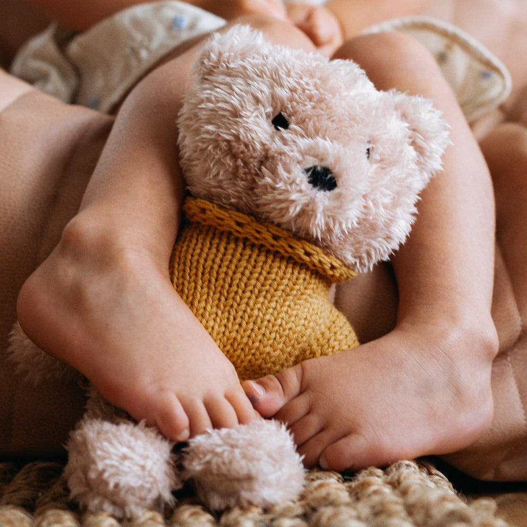 Dulcie Bear soft toy cuddled between a child’s feet, showing creamy vanilla plush fur and cozy mustard knitted jumper.