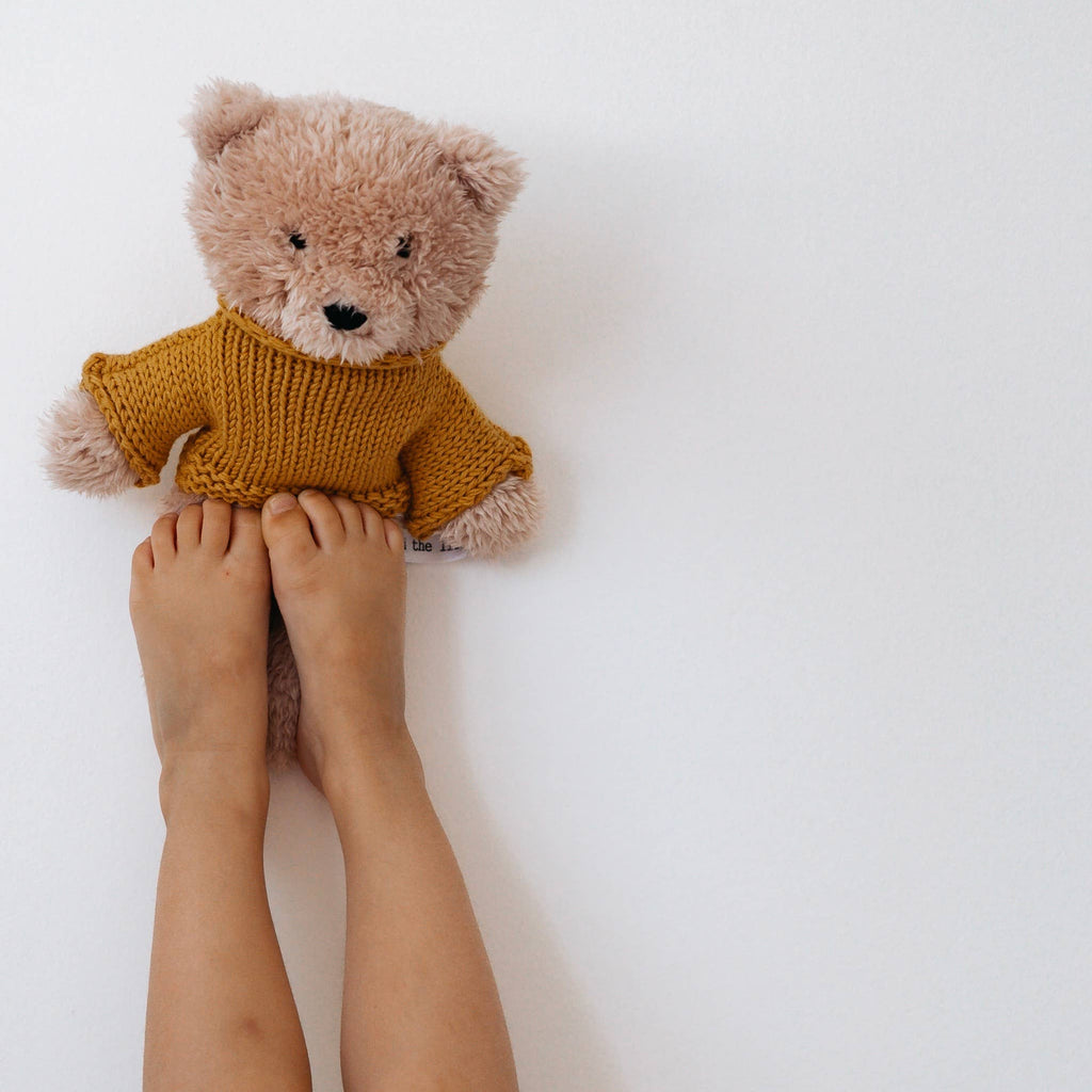 Child holding Dulcie Bear plush toy up by the feet against a white background, displaying her soft fur and knit jumper details.