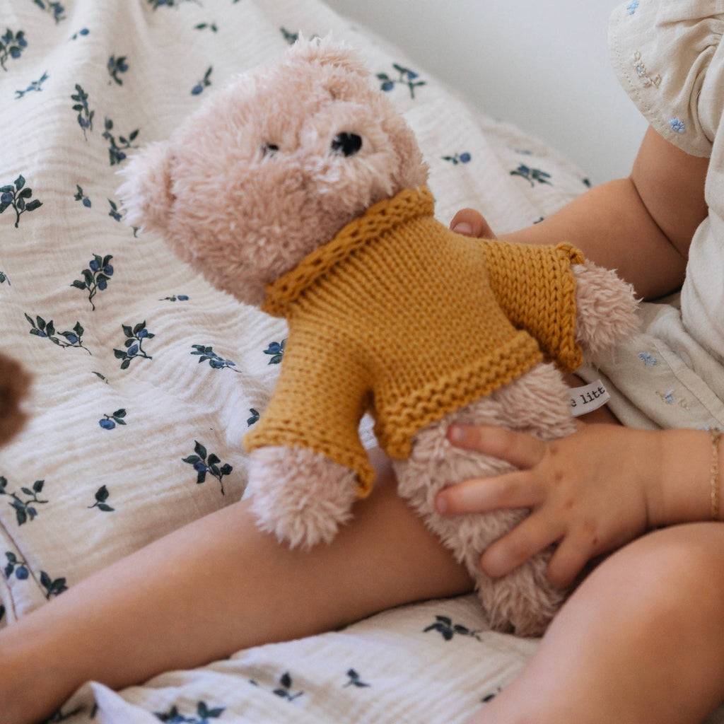 Child sitting in bed cuddling Dulcie Bear Mini, soft vanilla plush bear wearing mustard merino wool jumper, resting on floral bedding.