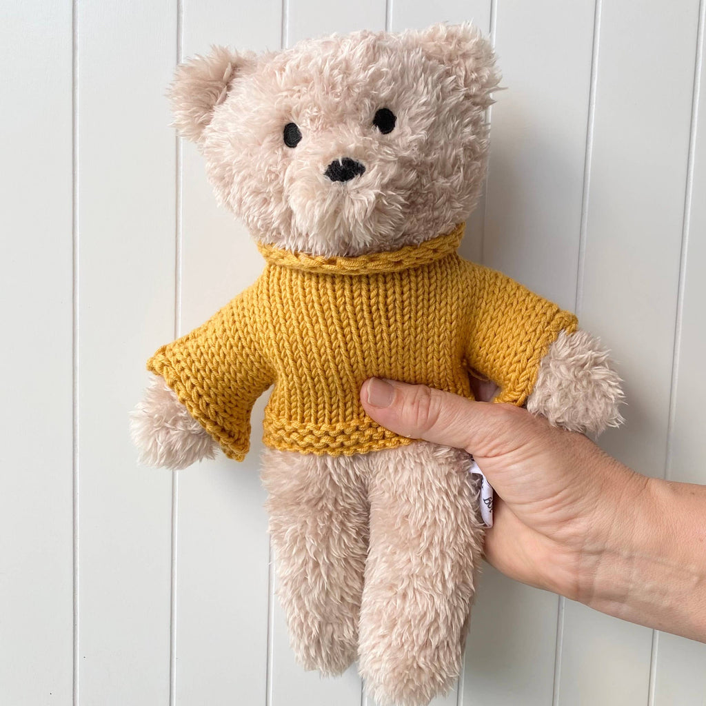 Hand holding Dulcie Bear Mini soft toy made from creamy vanilla plush fabric with mustard knitted merino wool jumper against white panel wall.