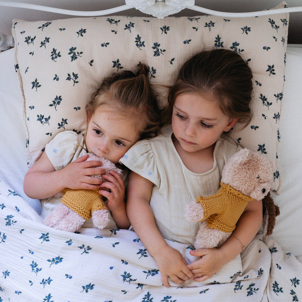 Two young girls lying in bed holding Dulcie Bear Mini soft toys, both bears made from vanilla plush with mustard knitted jumpers.
