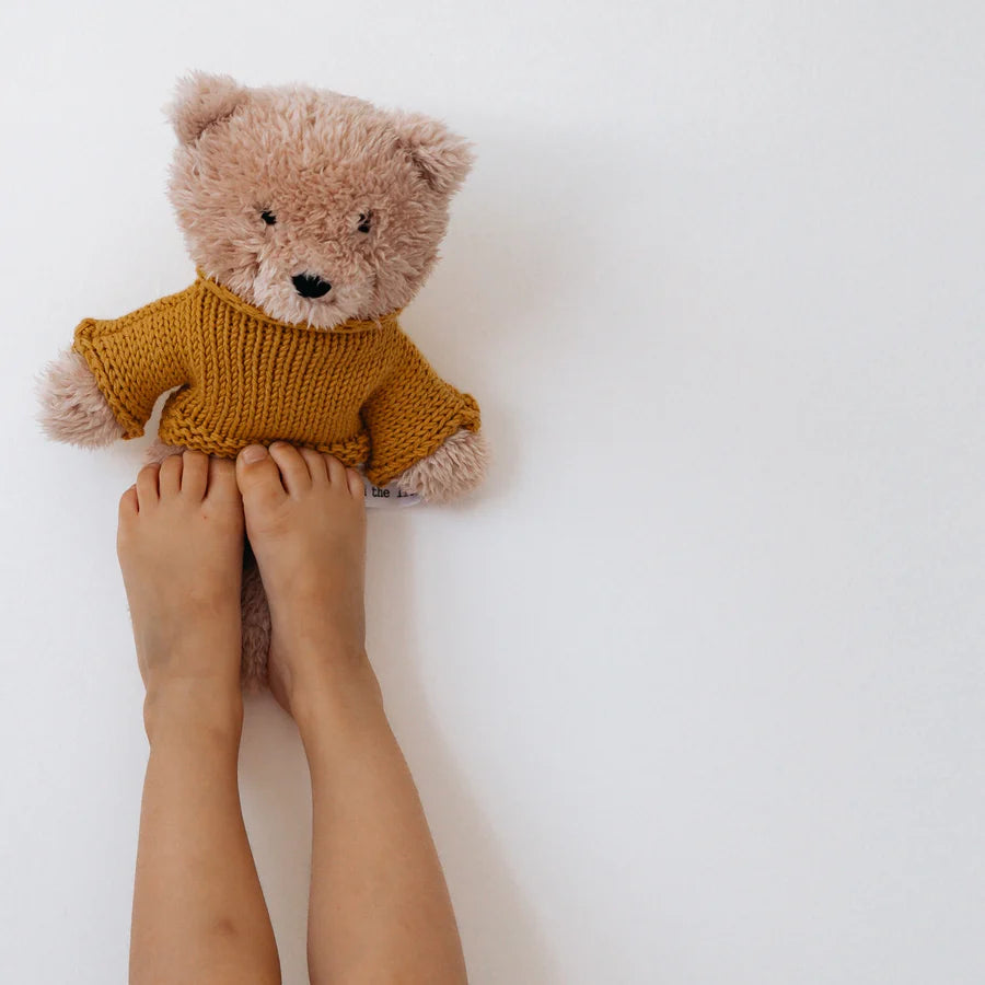Close-up of Dulcie Bear Mini soft toy with creamy vanilla plush and mustard merino wool jumper, placed between a child’s feet and white wall.