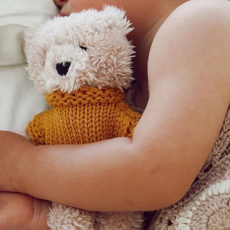 Baby cuddling Dulcie Bear Mini soft toy, creamy vanilla plush bear dressed in mustard merino wool jumper, resting against white blanket.