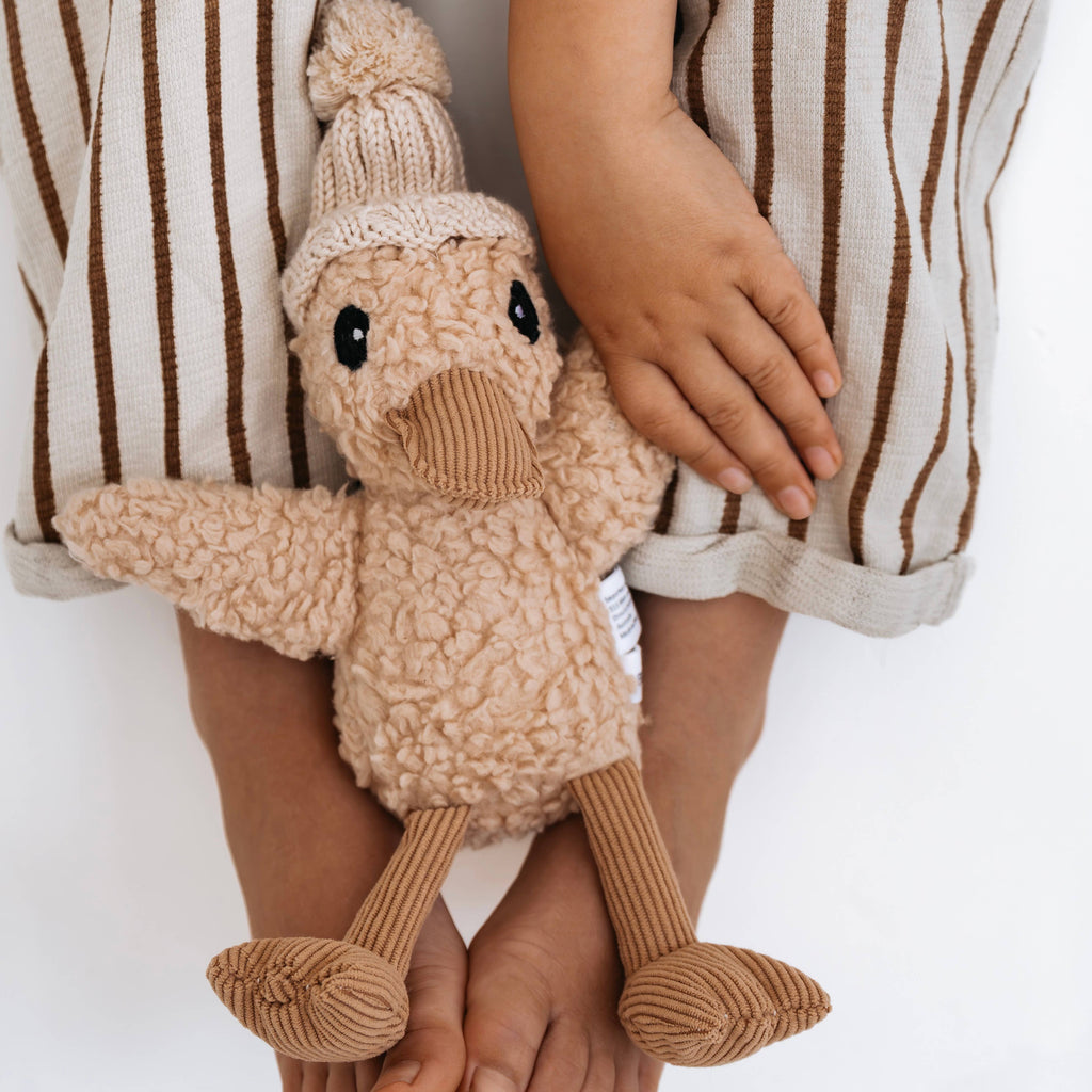Close-up of Edmond Duck soft toy being held by a child. Made from beige cotton plush with caramel corduroy beak and feet, wearing a cream merino wool knitted beanie.