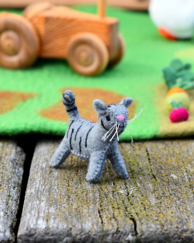 Handcrafted grey felt cat toy with black stripes and white whiskers, posed on a rustic wooden surface with colourful small world toys in the background.