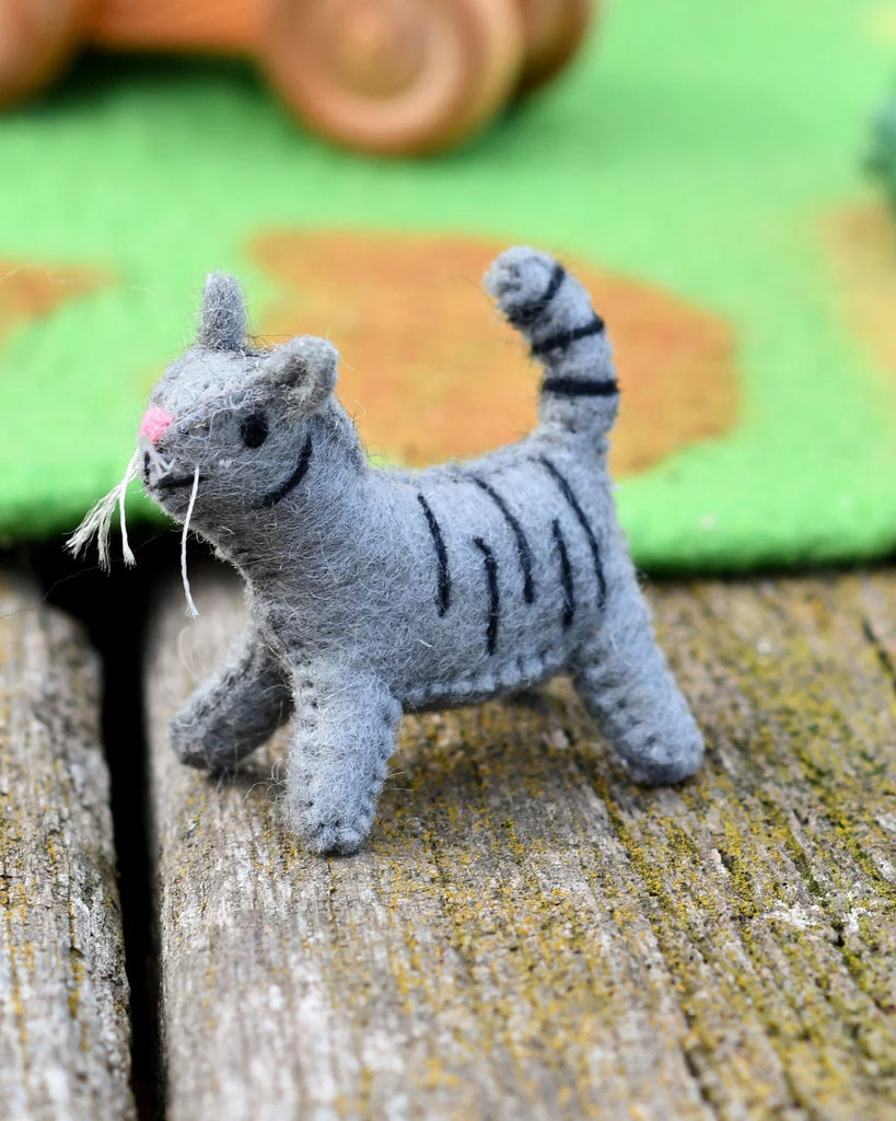 Side view of a grey felt cat toy with black stripes and stitched details, placed on a textured wooden background for small world imaginative play.