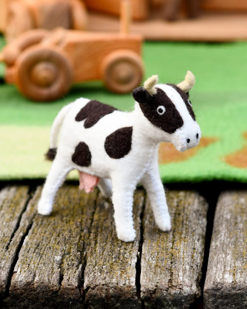 Side view of a handmade felt cow toy on a rustic wooden surface, surrounded by small world farm play props including wooden tractor wheels.
