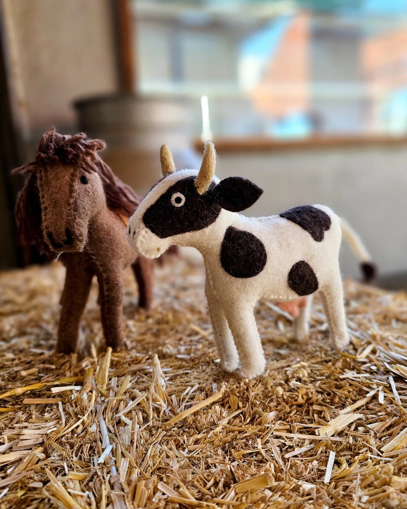 Felt cow toy placed in a straw-filled barnyard setup with a felt horse toy, surrounded by other felt farm animals for interactive storytelling play.