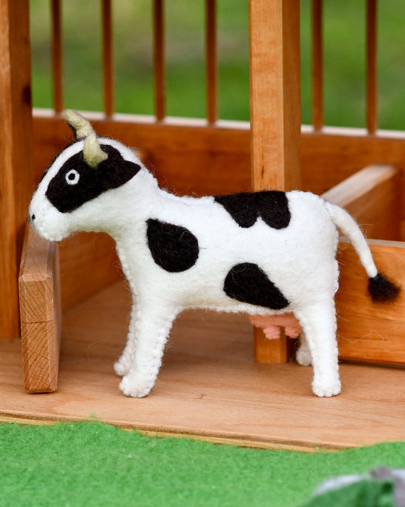Felt cow toy posed in front of a wooden farm fence on a green felt play mat, perfect for imaginative small world play.