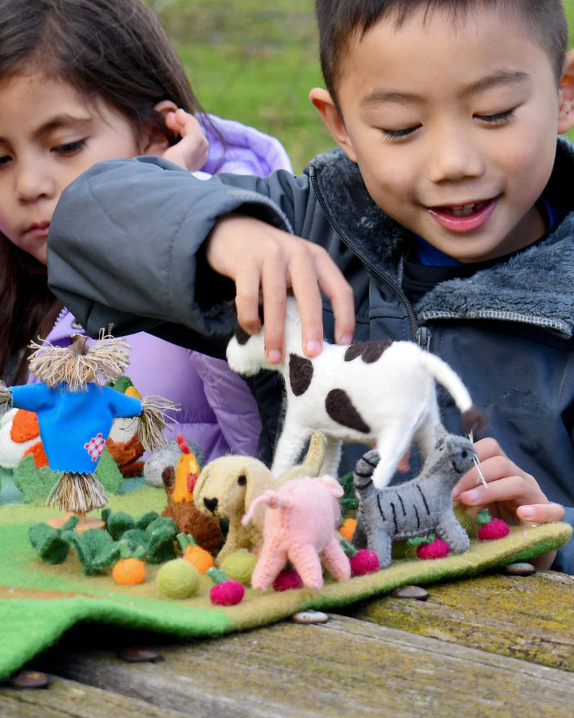 Group of children playing with a colourful set of felt farm animal toys, including the cow, at an outdoor play table.