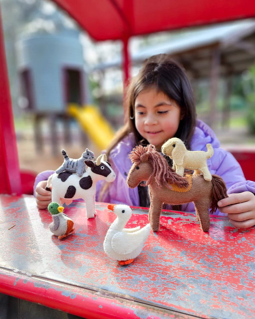 Smiling child playing with felt farm animals, including the cream felt dog, on a bright red outdoor table.