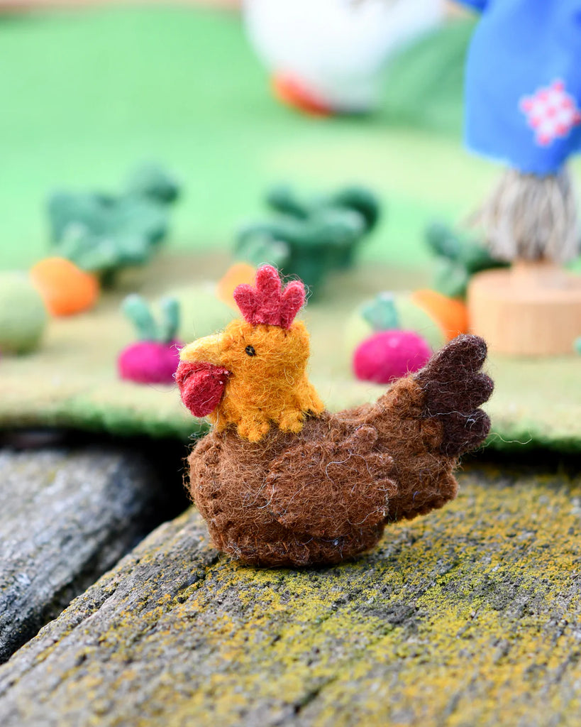 Side view of a brown felt chicken toy with red and yellow details, placed beside pink and yellow felt veggies in a small world farm play setup.