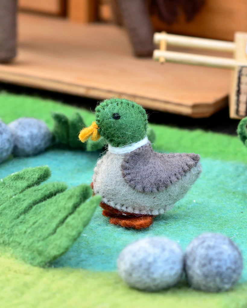 Felt duck toy standing near blue felt pebbles and a wooden toy stable, creating a playful farm pond scene.