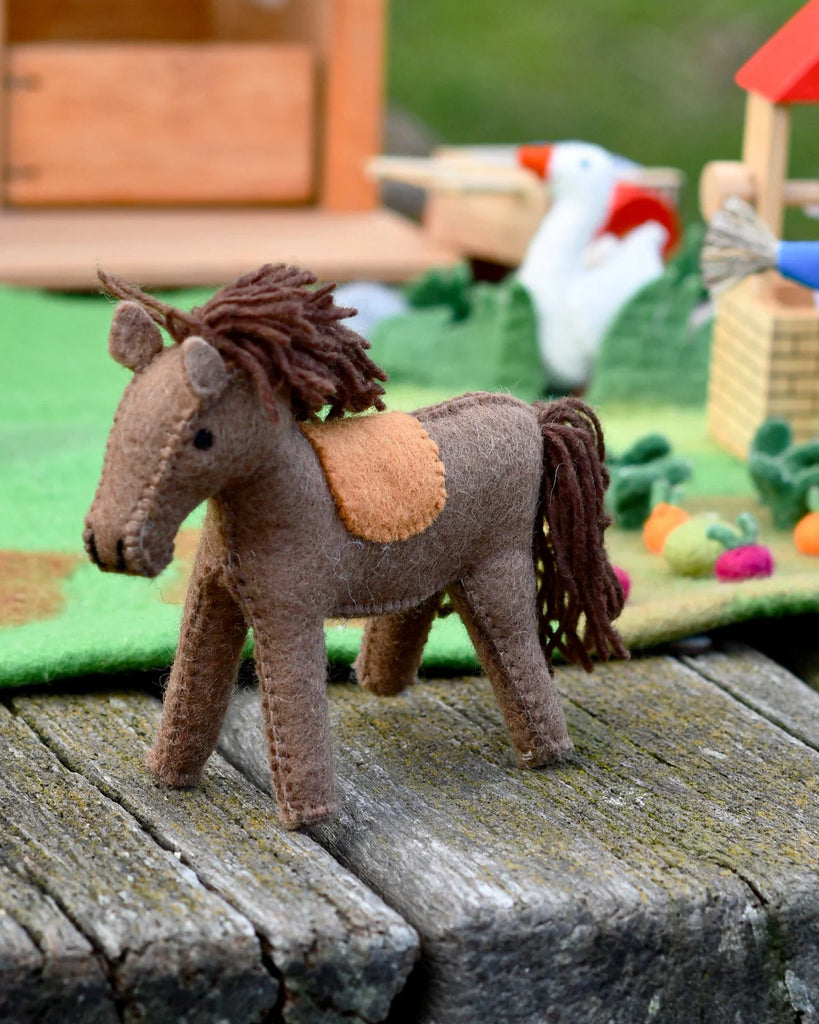 Side view of a felt horse toy with a wool mane, placed on a rustic wooden surface with a toy tractor in the background.
