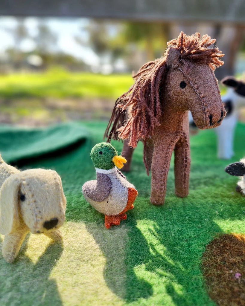 Brown felt horse toy on a green play mat surrounded by other felt farm animals, including a sheep and cow, in an outdoor play scene.