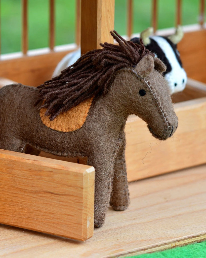 Handcrafted brown felt horse toy with white detailing, standing beside a wooden stable structure on a felt play surface.