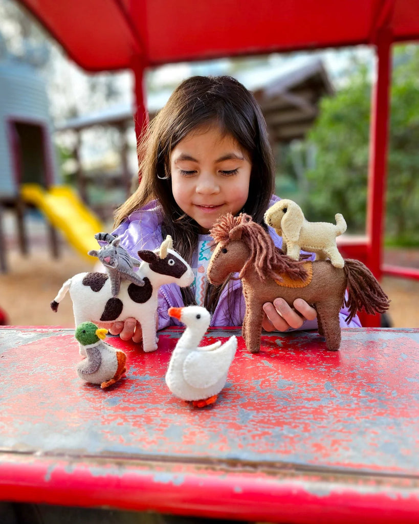 Smiling child holding a collection of handmade felt farm animals, including a brown felt horse, while seated at a red outdoor table.