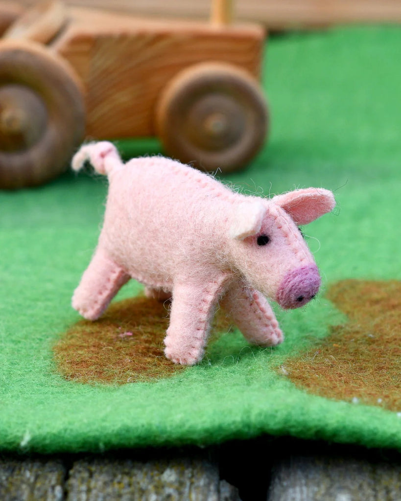 Side profile of a pink felt pig toy on a green felt base, next to a wooden toy tractor wheel, part of a small world farm setup.