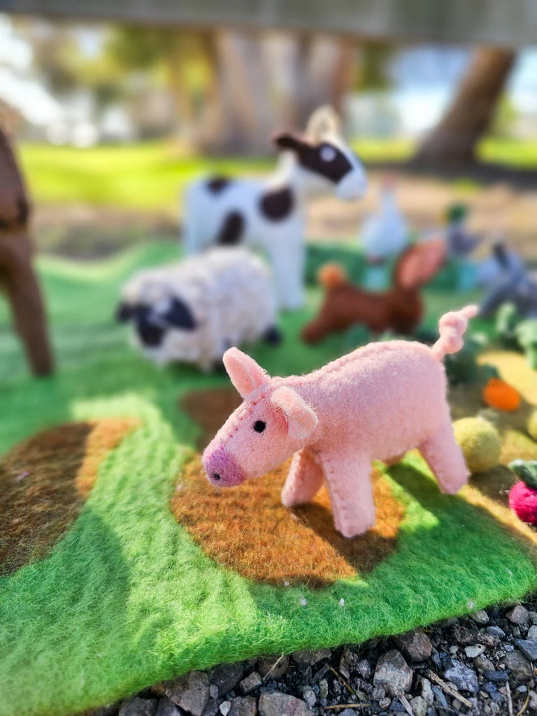 Felt pig toy surrounded by other handmade farm animals, set on a green felt farm landscape for creative pretend play.