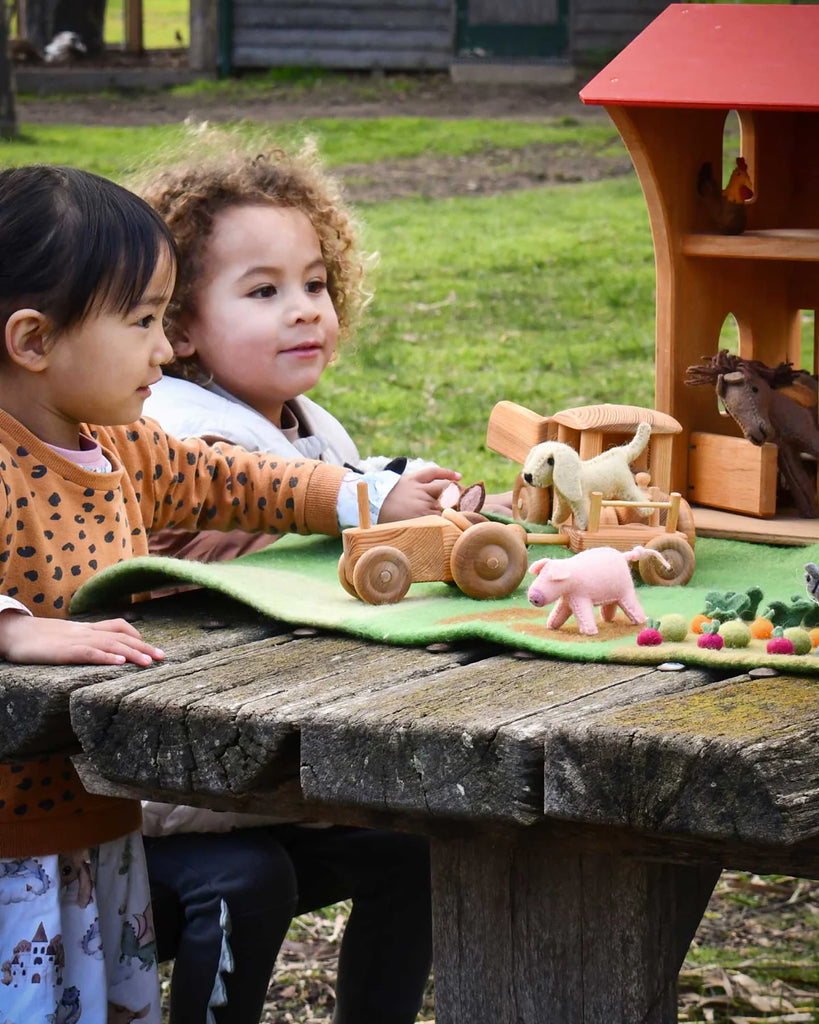 Two children playing outdoors with a set of handmade felt farm animal toys, including the pink pig, on a rustic wooden table.