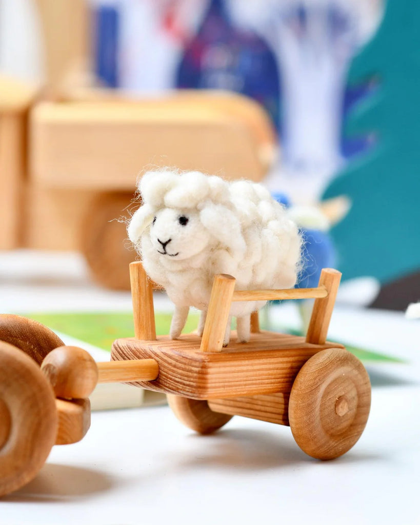 Fluffy white felt sheep toy sitting in a miniature wooden wagon, styled in a whimsical farm play scene for imaginative storytelling.