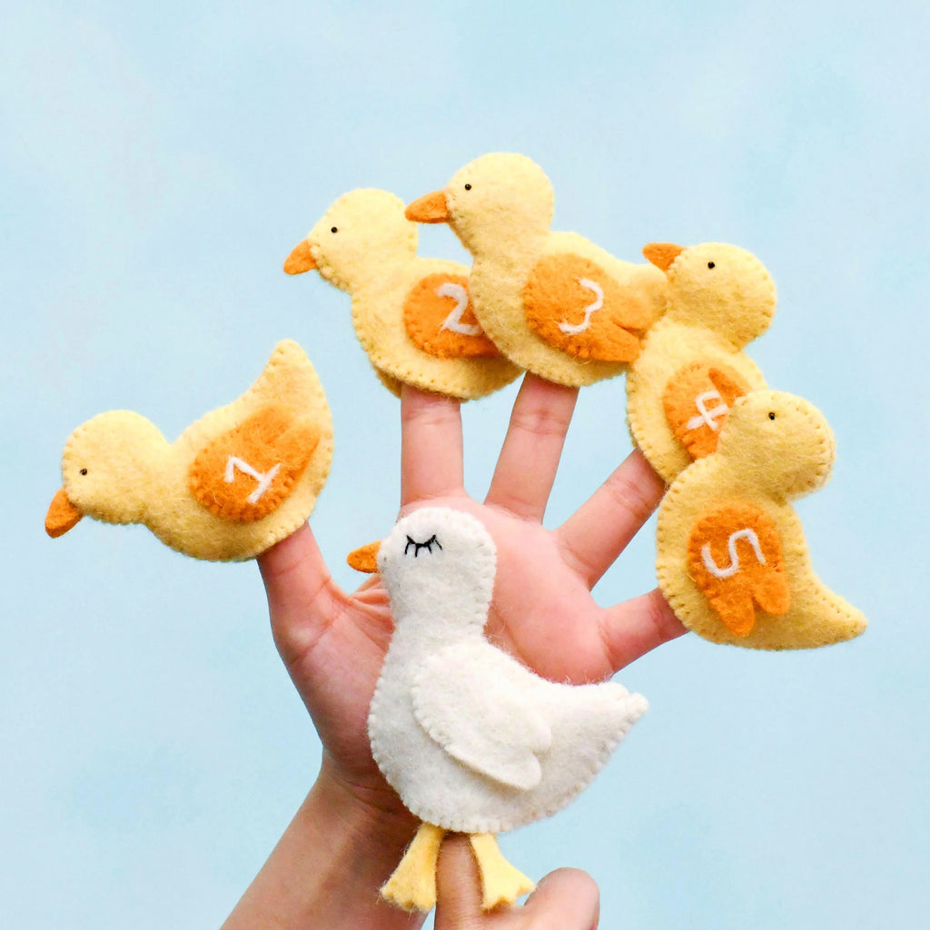 Hand holding five yellow felt duckling finger puppets, each with a number from 1 to 5 stitched on their wings.