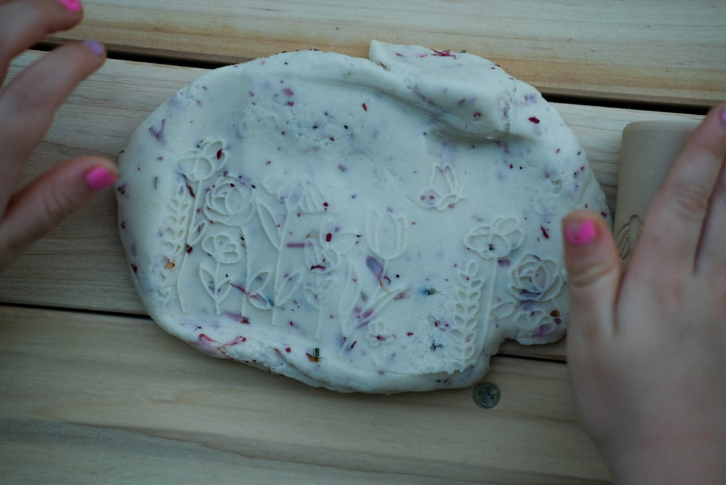 Top-down view of flower-printed white playdough with embedded dried petals, rolled using biodegradable floral dough roller.