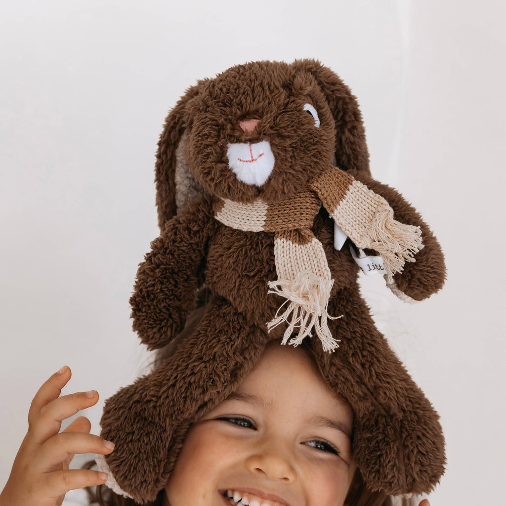 Medium Frankie Rabbit soft toy made from chocolate brown plush fabric with long ears, wearing a knitted merino wool scarf, held above a smiling child’s head.
