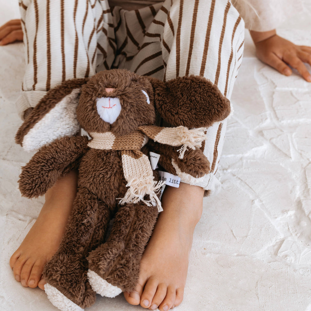Child sitting cross-legged holding Frankie Rabbit Medium, chocolate brown plush bunny with merino wool scarf, resting on white textured bedding.