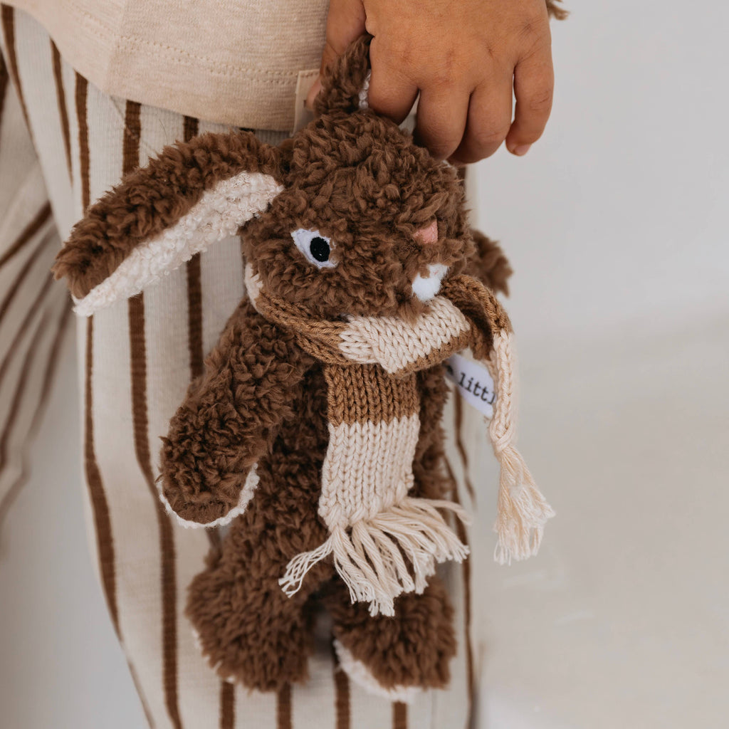 Close-up of Frankie Rabbit mini soft toy with brown plush fur, floppy ears, and a cream merino wool scarf, held by a child wearing striped pants.