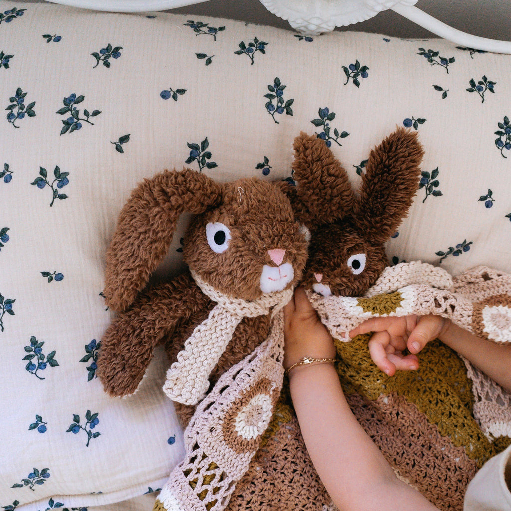 Two plush rabbits, 1 medium and 1 mini in size, Frankie and a friend, lying on a floral bedsheet, both with floppy ears and cozy knitted accessories, next to a child’s hands.