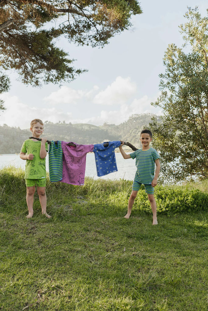 Two children standing outdoors with colorful clothes hanging on a line, with trees and water in the background.