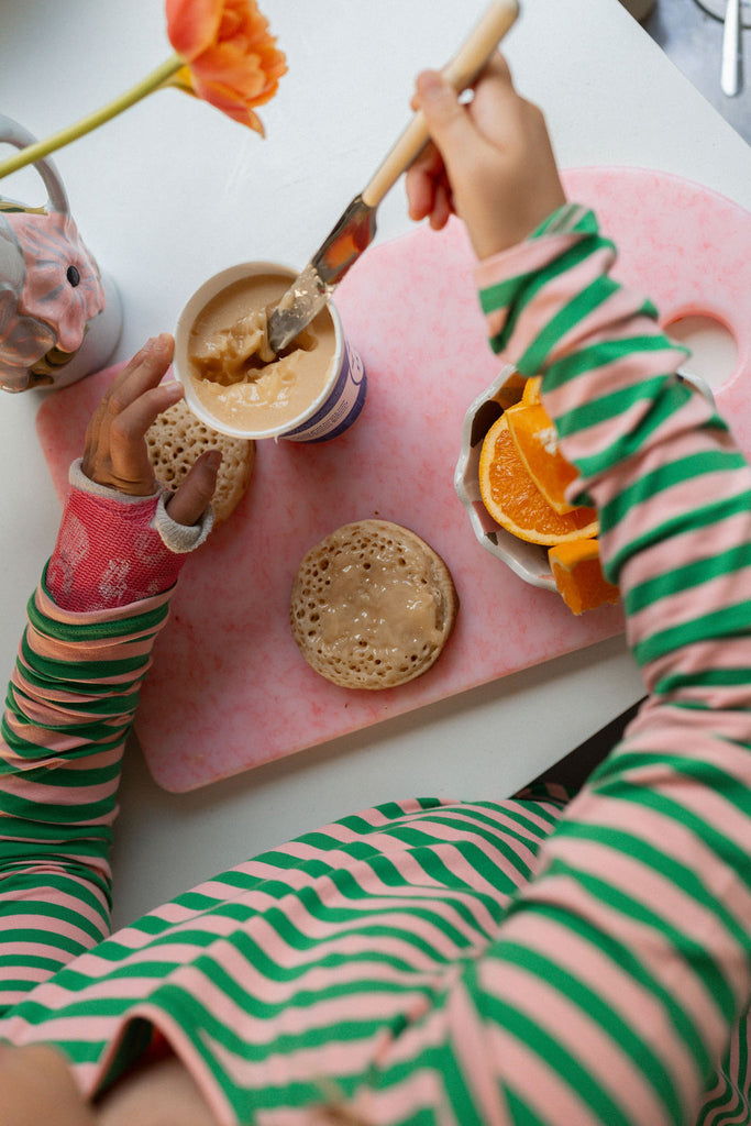 Child wearing G.Nancy bubblegum and green striped PJs sitting at a breakfast table, spreading peanut butter on toast with fresh orange juice nearby.
