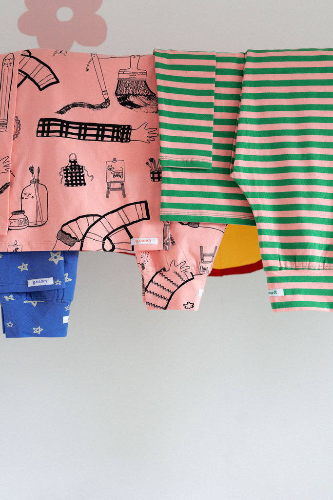 Assortment of colourful G.Nancy kids’ pyjamas, including pink and green striped set, hanging on a clothesline above an art-filled craft table.