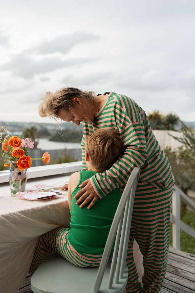 Parent hugging child wearing G.Nancy striped PJs while sitting on a veranda with flowers and breakfast setup, ocean view in the background.
