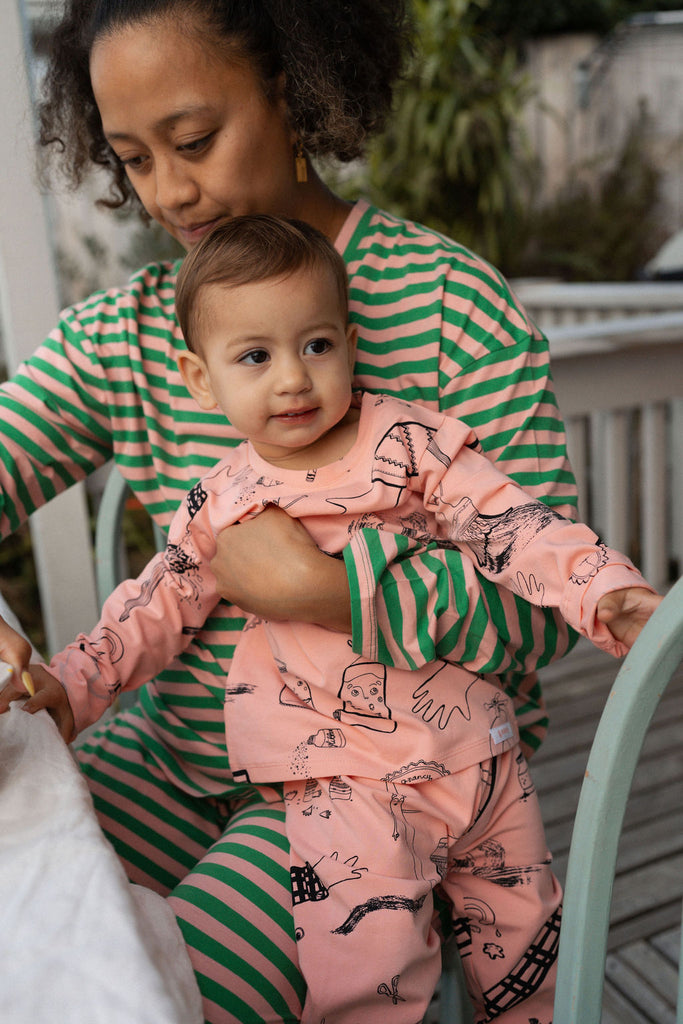 Mother in G.Nancy green and pink striped women’s pajamas holding a baby in pink illustrated pajamas, both smiling on a porch.