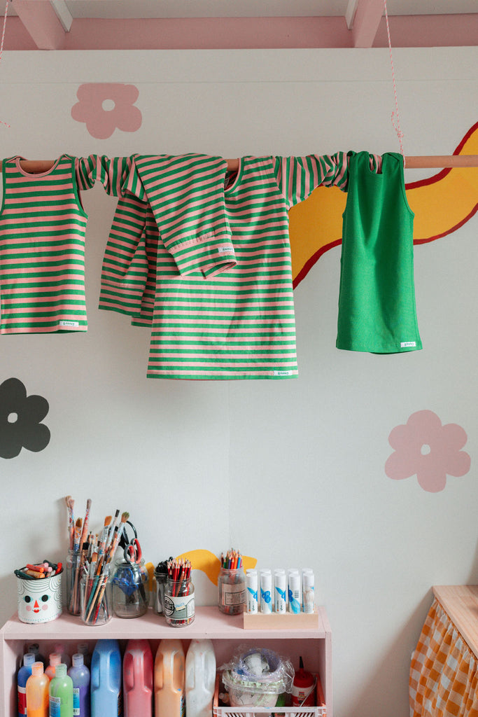 G.Nancy bubblegum and kelly green striped pajama top and pants hanging on a playful pastel children’s room clothesline surrounded by art supplies.