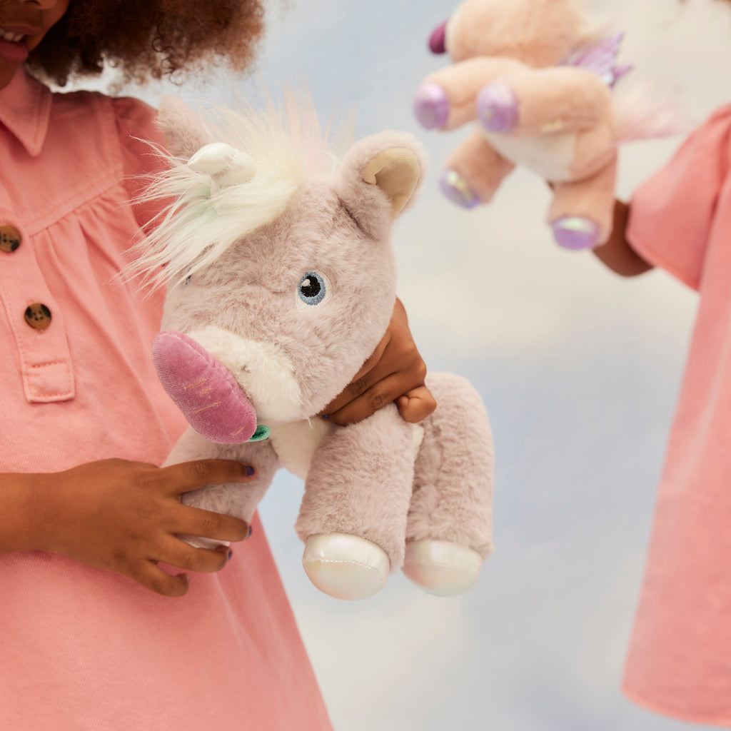 Child holding Glimmer unicorn plush – Smiling girl in pink shirt cuddling lilac unicorn with iridescent details.