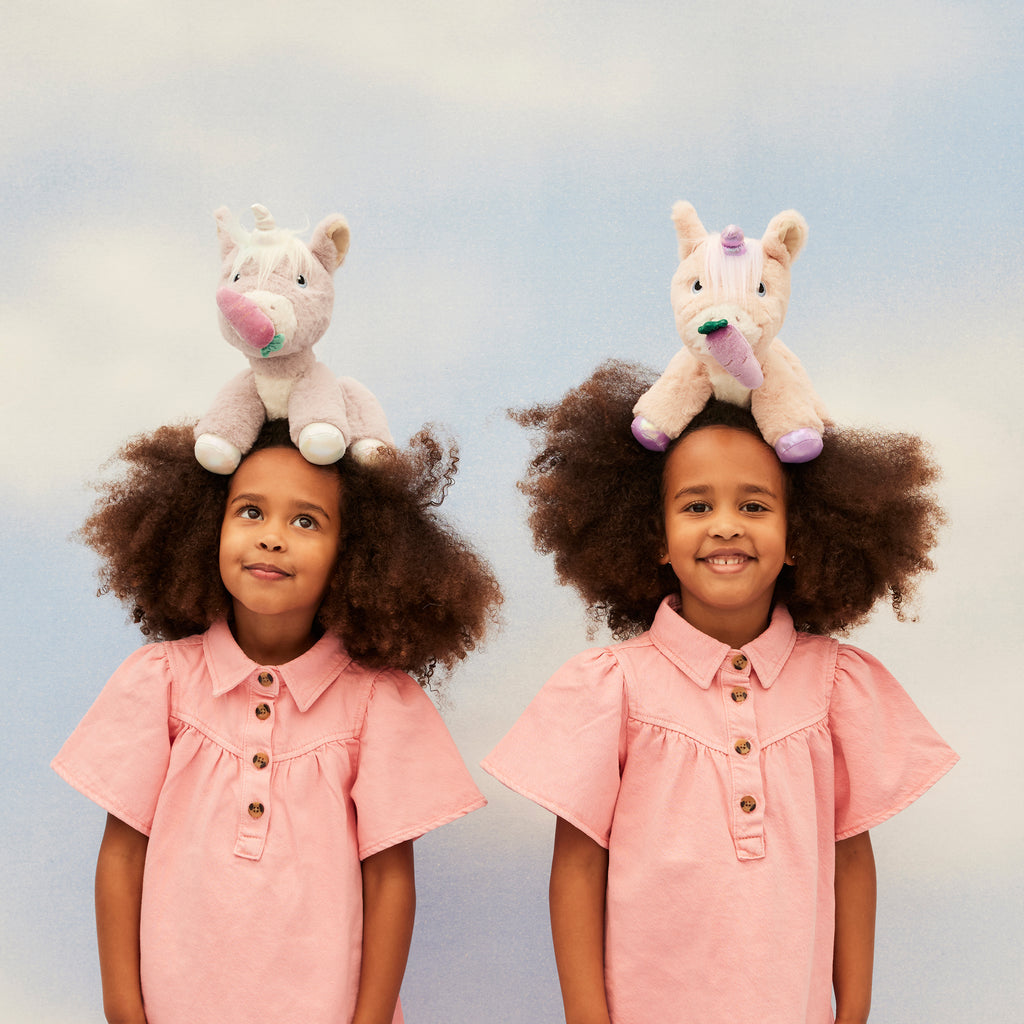 Two children balancing Glimmer and Shimmer unicorn plush toys on heads – Playful moment against soft blue background.