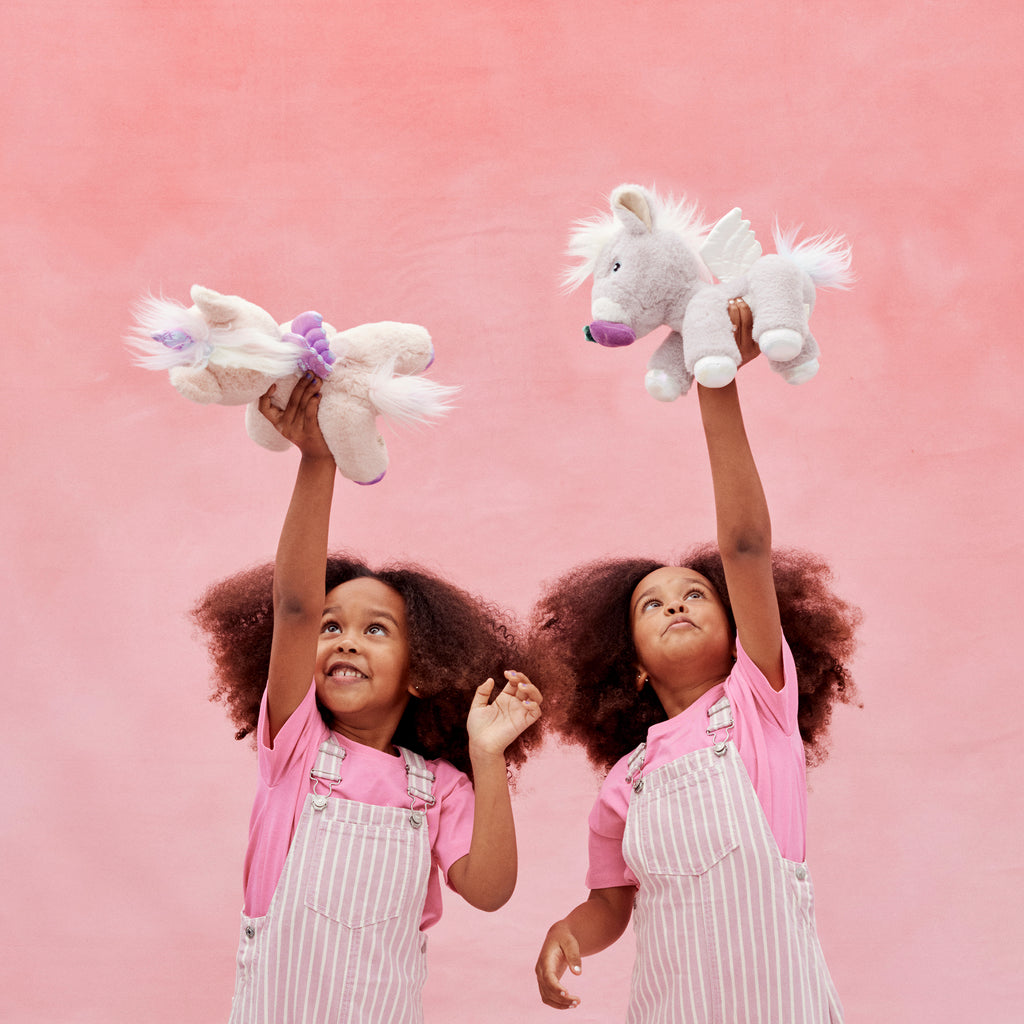 Children raising unicorn plush toys in the air – Joyful scene with lilac and pink Dinkum Unicorns in a pink backdrop.
