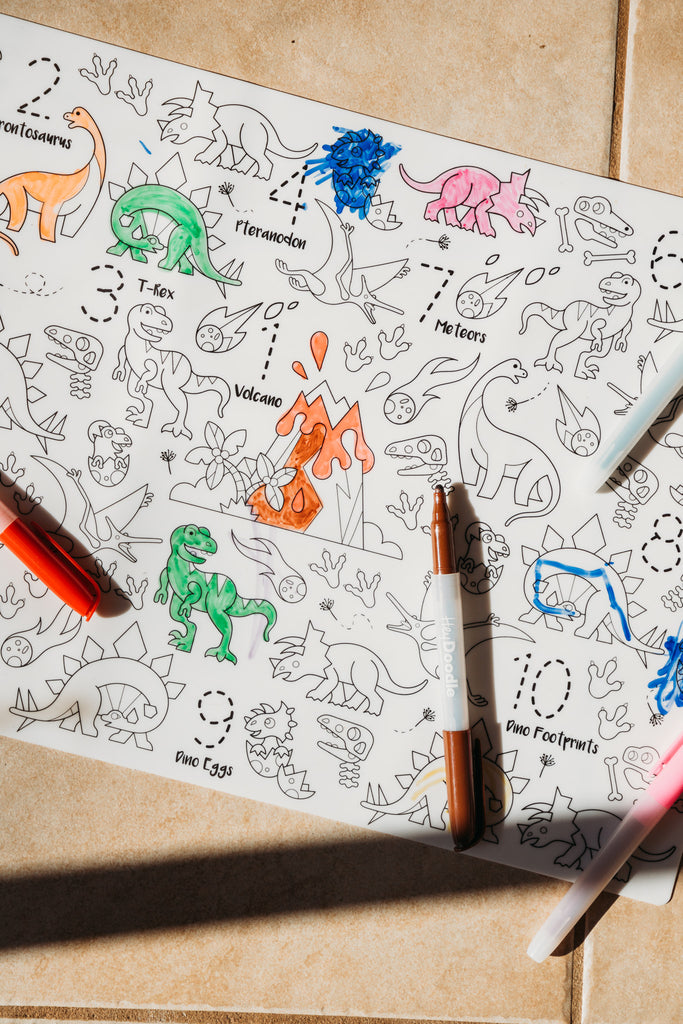 Coloring silicone mat with dinosaur illustrations and coloring markers on a beige surface