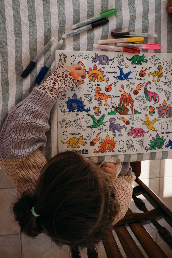 Child coloring on a Hey Doodle Silicone Mat with variously colored dinosaurs on a striped fabric background.