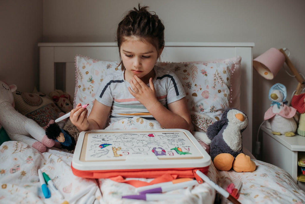 Child colouring the HeyDoodle Into the Wild mat on a bed, surrounded by toys.