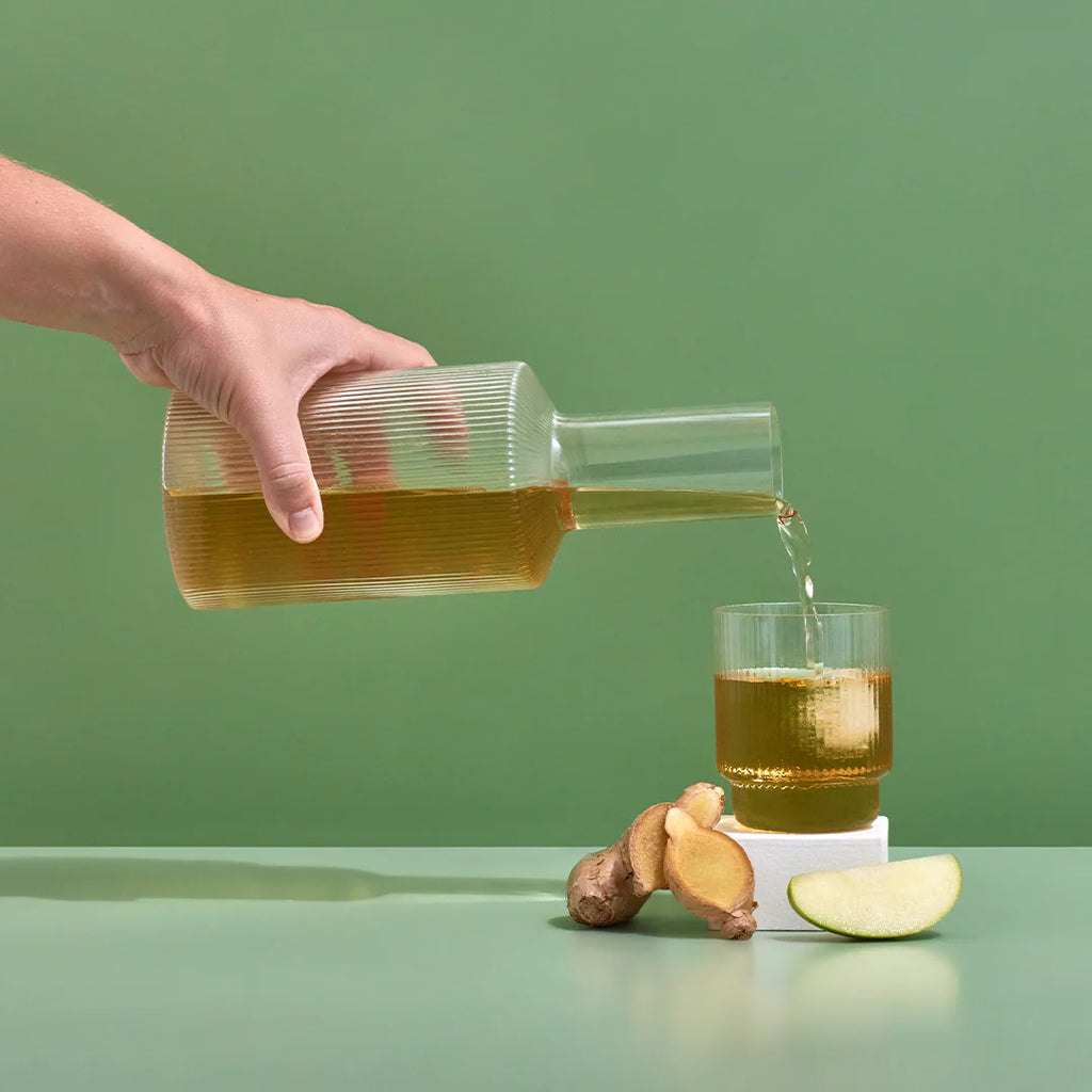 Hand pouring Hydramama Apple & Ginger drink from a glass carafe into a tumbler, styled with ginger, lemon, and apple slices.