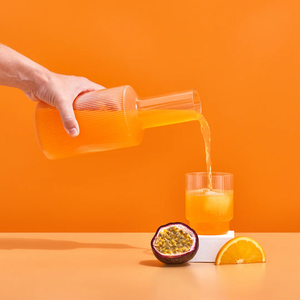 Hydramama Passionfruit & Orange Hydration Electrolyte Drink for Pregnancy & Postpartum - Hydramama being poured from jug into glass with ice. pictured nearby is a fresh passionfruit cut in half and a slice of fresh orange.