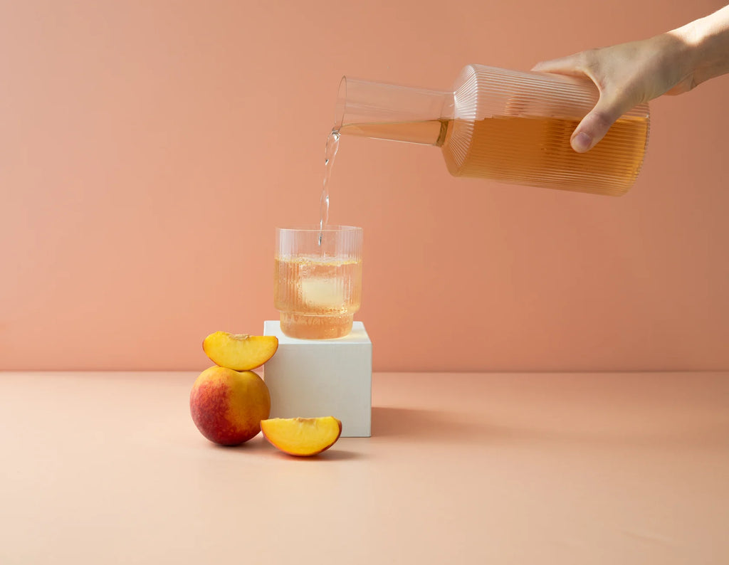 Hydramama Peach Iced Tea Hydration Electrolyte Drink for Pregnancy & Postpartum - Pouring drink from jug into glass with ice fresh peaches sitting nearby
